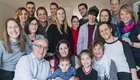 Eine große Gruppe von Familienmitgliedern versammelt sich in einem hellen Raum. Im Vordergrund stehen zwei ältere Erwachsene, während mehrere jüngere Personen fröhlich im Hintergrund lächeln. Alle sind eng beieinander und strahlen Zuversicht und Gemeinsamkeit aus.