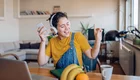 Eine junge Frau sitzt am Tisch. Sie hat Kopfhörer auf und das Handy in ihrer rechten Hand. Sie hört Musik, singt mit und bewegt sich im Takt. Das Bild verbreitet gute Laune.