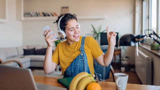 Eine junge Frau sitzt am Tisch. Sie hat Kopfhörer auf und das Handy in ihrer rechten Hand. Sie hört Musik, singt mit und bewegt sich im Takt. Das Bild verbreitet gute Laune.