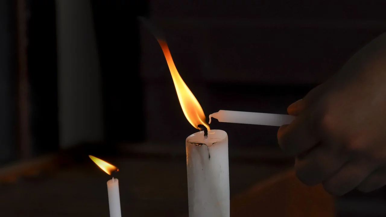 Eine Hand hält eine unbenutzte Kerze, während sie eine brennende Kerze anzündet. Die Flamme der brennenden Kerze leuchtet im Dunkeln. Im Bild sind zwei Kerzen sichtbar, eine ist bereits angezündet.