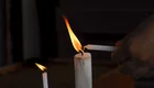 Eine Hand hält eine unbenutzte Kerze, während sie eine brennende Kerze anzündet. Die Flamme der brennenden Kerze leuchtet im Dunkeln. Im Bild sind zwei Kerzen sichtbar, eine ist bereits angezündet.