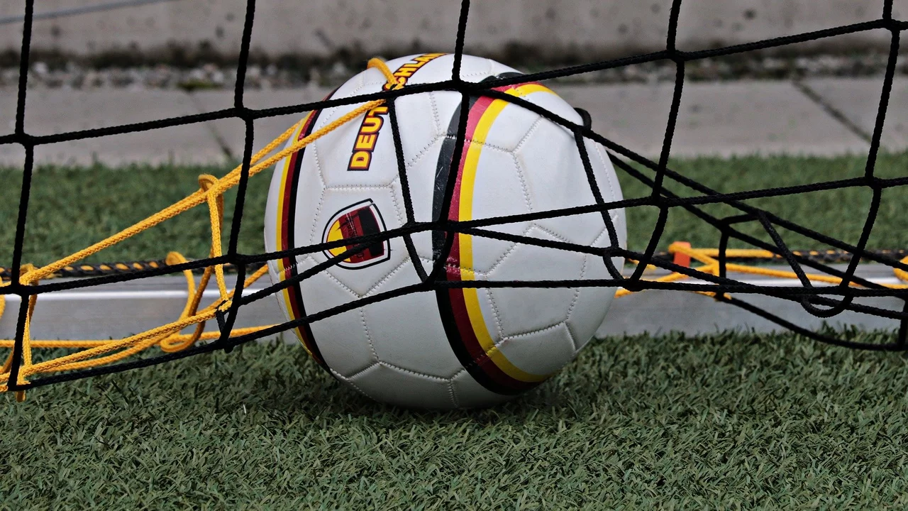 Ein Fußball liegt am Boden, teilweise im Netz eines Torwarts. Die obere Hälfte des Balls zeigt die deutschen Farben Schwarz, Rot und Gold. Der Hintergrund besteht aus grünem Kunstrasen.