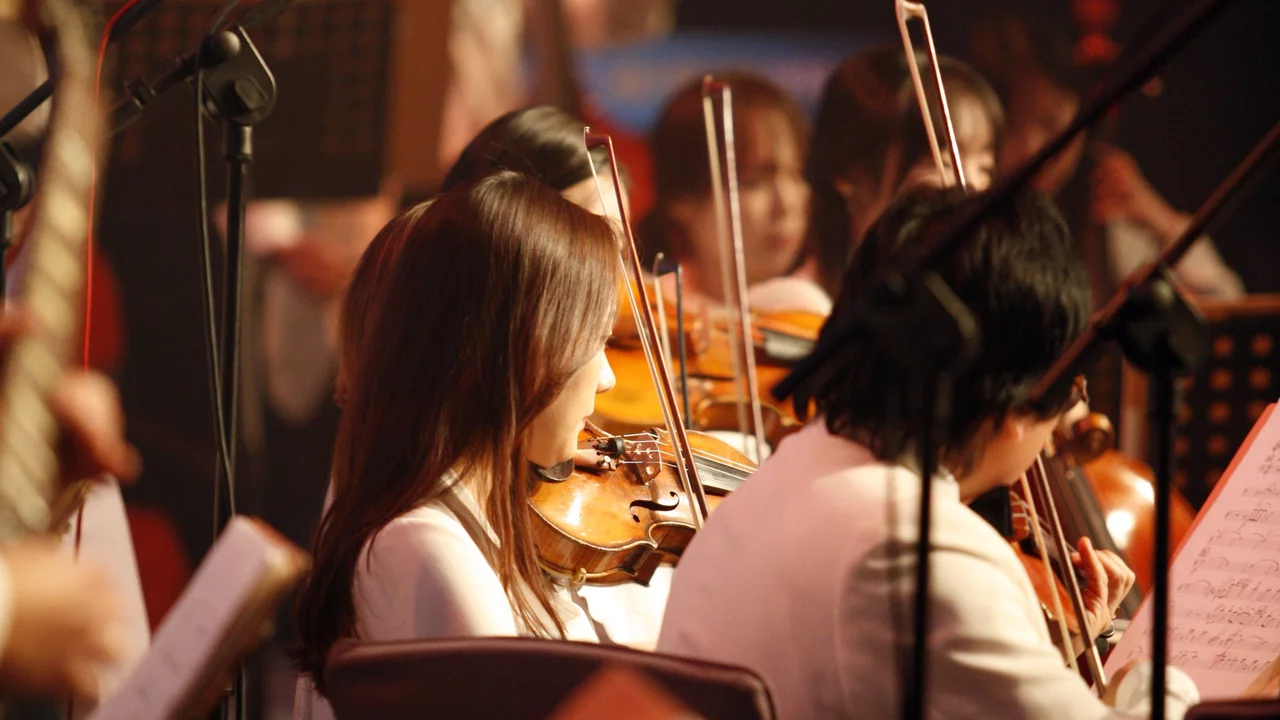 Eine Gruppe von Musikern spielt in einem Orchestersetting. Im Vordergrund stehen Violinspieler, die konzentriert musizieren. Das Licht ist sanft, und Notenblätter sind sichtbar. Die Atmosphäre vermittelt eine Leidenschaft für die Musik.