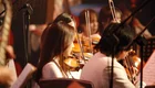 Eine Gruppe von Musikern spielt in einem Orchestersetting. Im Vordergrund stehen Violinspieler, die konzentriert musizieren. Das Licht ist sanft, und Notenblätter sind sichtbar. Die Atmosphäre vermittelt eine Leidenschaft für die Musik.