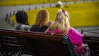 Drei Frauen sitzen auf einer Bank in einem Park, alle von hinten sichtbar. Die Umgebung ist grün mit Bäumen und Wegen. Die Frauen tragen unterschiedliche Frisuren und Kleidung, während sie gemeinsam die Aussicht genießen.