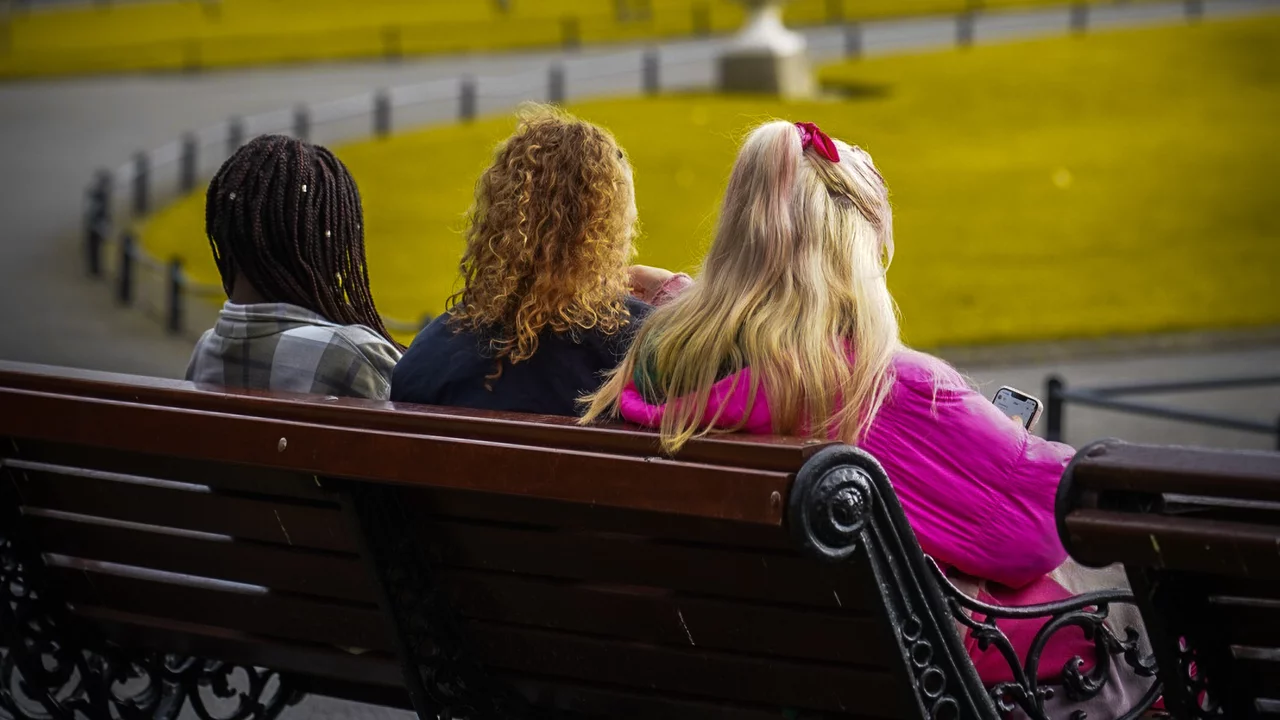 Drei Frauen sitzen auf einer Bank in einem Park, alle von hinten sichtbar. Die Umgebung ist grün mit Bäumen und Wegen. Die Frauen tragen unterschiedliche Frisuren und Kleidung, während sie gemeinsam die Aussicht genießen.