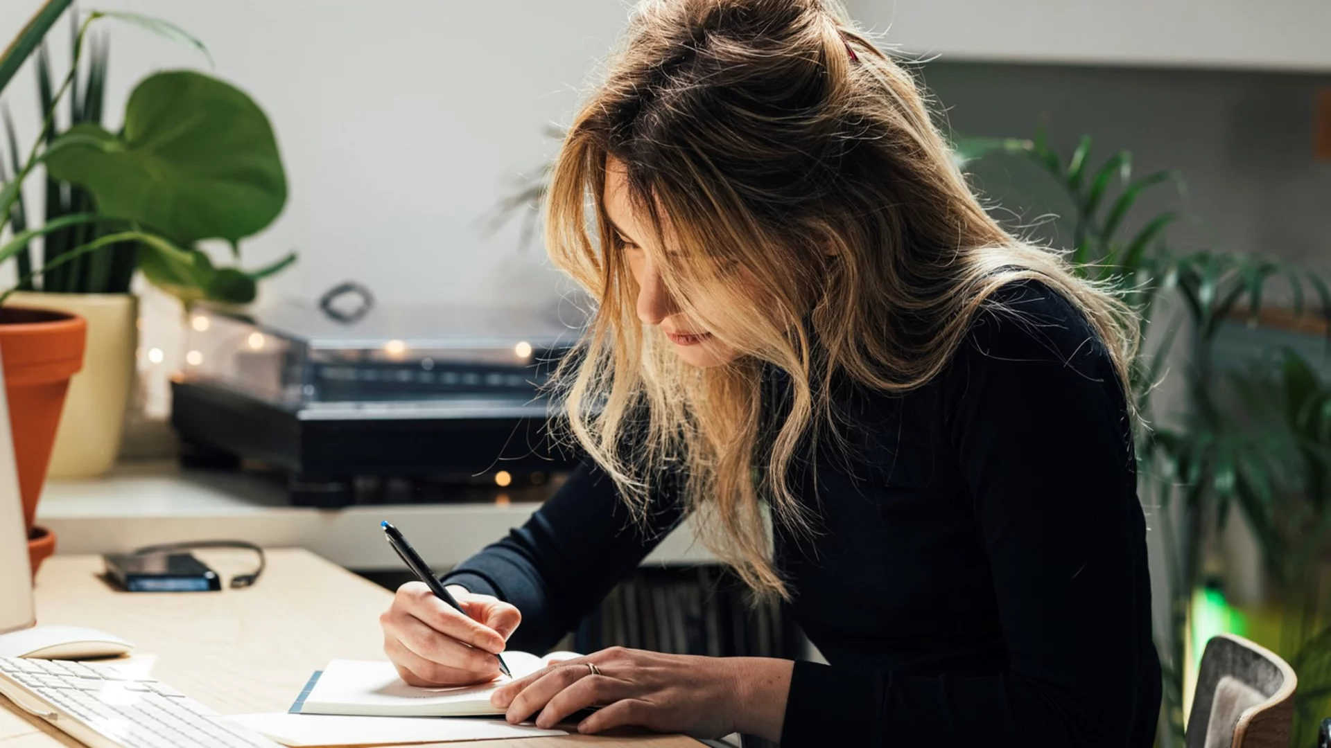 Eine Frau mit langen, blonden Haaren sitzt an einem Tisch und schreibt in ein Notizbuch. Sie trägt ein schwarzes Oberteil und ist konzentriert. Im Hintergrund sind Pflanzen und ein Schreibtisch mit einem Computer sichtbar.