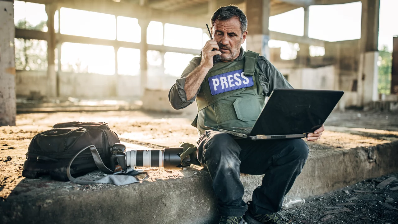 Ein Journalist in Schutzweste mit der Aufschrift "PRESS" sitzt auf einem Betonblock. Er spricht am Telefon und arbeitet am Laptop. Neben ihm liegt eine Kameratasche mit einer Kamera und Objektiven. Im Hintergrund sind verlassene, industrielle Ruinen zu sehen.