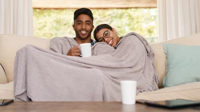 Ein lächelndes Paar sitzt gemütlich auf einem Sofa, eingekuschelt in eine Decke. Sie halten Tassen in den Händen und genießen gemeinsam eine entspannte Zeit. Im Hintergrund sind helle Vorhänge und ein grüner Ausblick zu sehen.