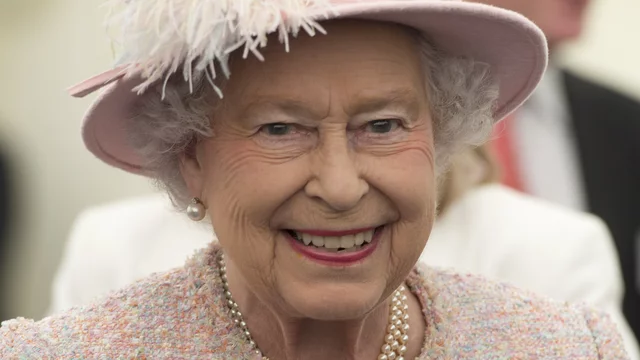 Eine lächelnde Frau mit einer auffälligen rosa Kopfbedeckung und einem hellen, strukturierten Ensemble. Sie trägt Perlenohrringe und einen passenden Perlenschmuck. Der Hintergrund ist unscharf und zeigt mehrere verschwommene Personen. Es ist Elizabeth II.
