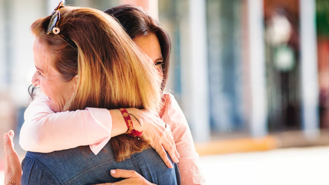 Zwei Frauen umarmen sich herzlich, lächelnd in einer angenehmen Umgebung. Die Szene strahlt Freude und Verbundenheit aus. Eine der Frauen trägt eine blaue Jeansjacke, während die andere in einem hellen Oberteil gekleidet ist.