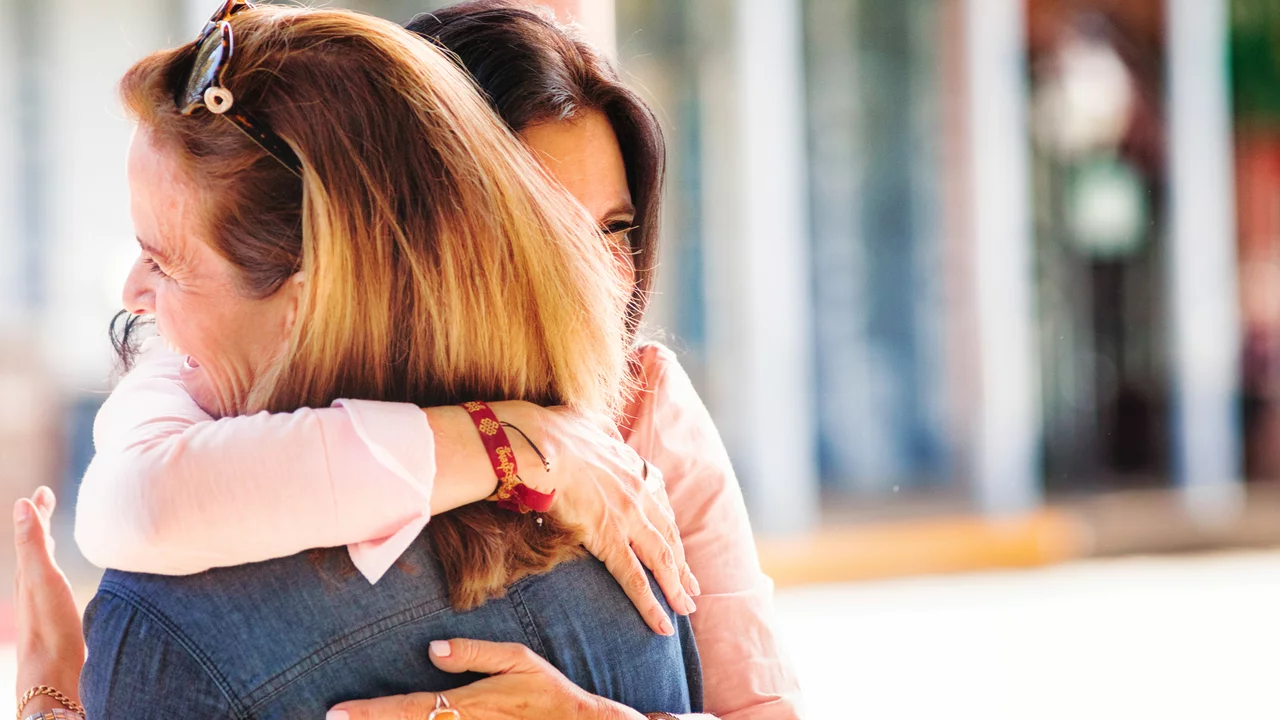 Zwei Frauen umarmen sich herzlich, lächelnd in einer angenehmen Umgebung. Die Szene strahlt Freude und Verbundenheit aus. Eine der Frauen trägt eine blaue Jeansjacke, während die andere in einem hellen Oberteil gekleidet ist.