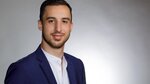 Ein junger Mann mit kurzem, dunklem Haar und einem leichten Bart lächelt freundlich. Er trägt einen blauen Anzug und ein weißes Hemd. Der Hintergrund ist hell und neutral, was den Fokus auf sein Gesicht legt.