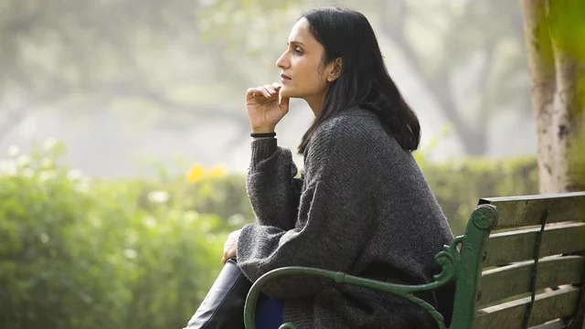 Frau sitzt nachdenklich auf Parkbank, schaut in die Ferne und hat die rechte Hand ans Kinn gelehnt