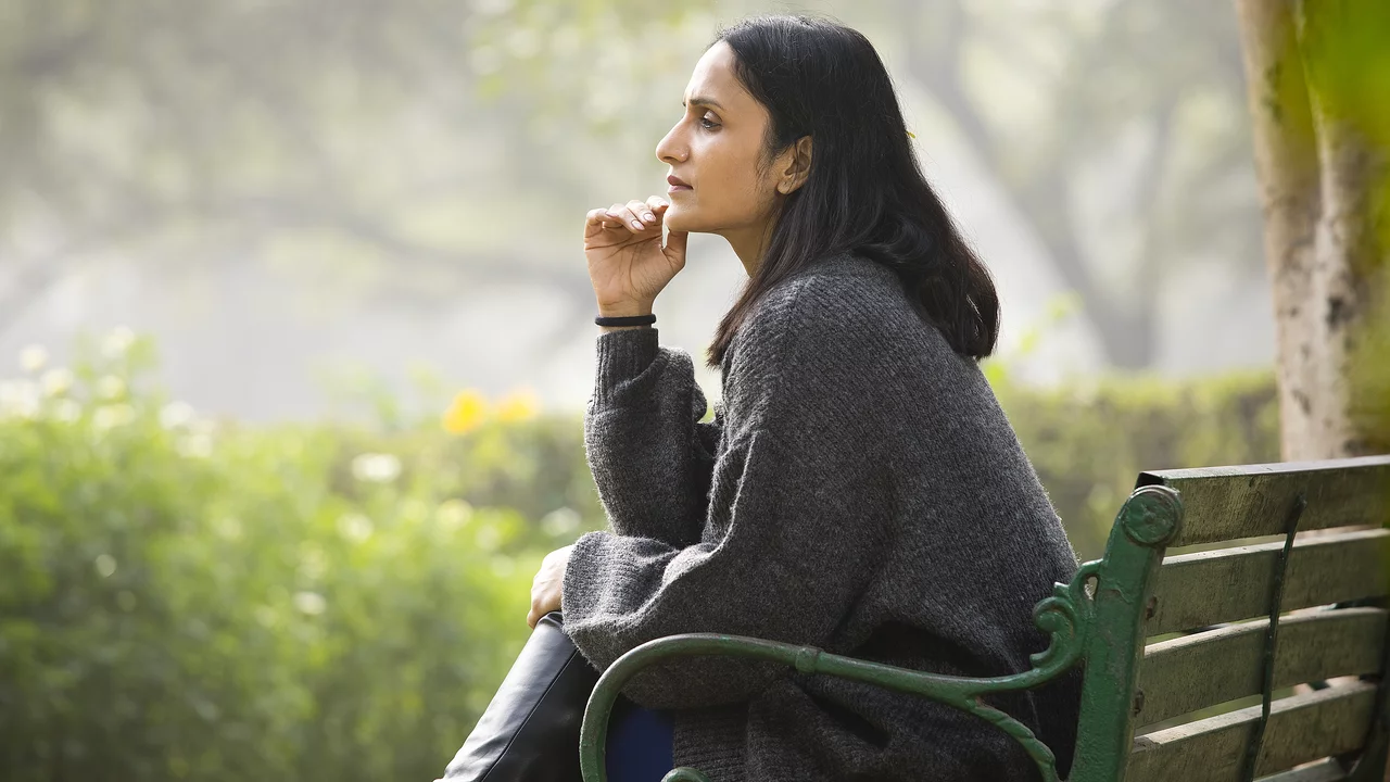 Frau sitzt nachdenklich auf Parkbank, schaut in die Ferne und hat die rechte Hand ans Kinn gelehnt