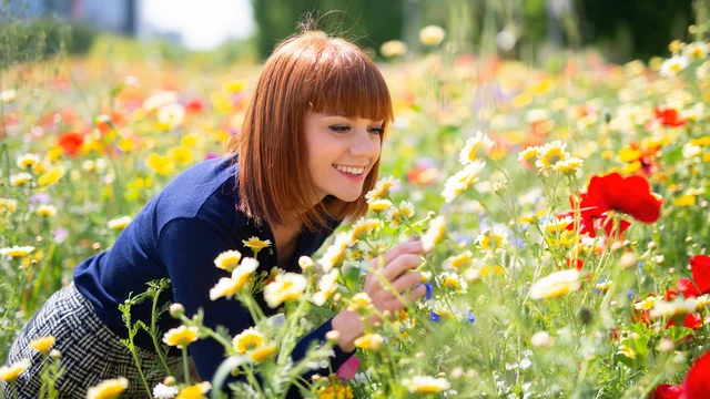 Eine lächelnde, lockige Frau mit rotem Haar beugt sich über ein buntes Blumenfeld, das mit verschiedenen Blumen in Gelb, Rot und anderen Farben blüht. Sie scheint die Blumen zu betrachten oder zu riechen und strahlt Freude aus.