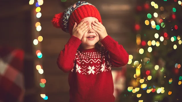Ein fröhliches Kind in einem roten Pullover mit Weihnachtsmuster und einer Mütze hält sich die Hände vor die Augen. Im Hintergrund sind verschwommene Lichter und ein weihnachtlicher Baum zu erkennen. Das Kind lächelt, als ob es auf eine Überraschung wartet.