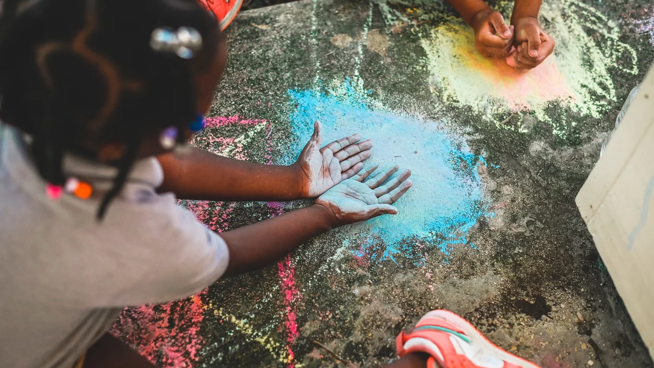 Zwei Kinder sitzen auf dem Boden und zeichnen mit bunter Kreide. Ein Kind hat die Hände auf den Boden gelegt, während das andere mit Kreide symbolische Formen um die Hände malt. Die Atmosphäre ist kreativ und verspielt, mit bunten Farbtönen auf dem Beton.