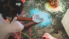 Zwei Kinder sitzen auf dem Boden und zeichnen mit bunter Kreide. Ein Kind hat die Hände auf den Boden gelegt, während das andere mit Kreide symbolische Formen um die Hände malt. Die Atmosphäre ist kreativ und verspielt, mit bunten Farbtönen auf dem Beton.