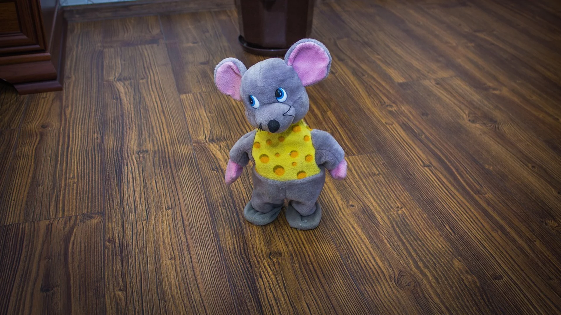 Ein plüschige Maus steht auf dem Boden. Sie trägt ein gelbes Oberteil mit Käse-Muster und hat große Ohren mit rosa Innenseiten. Der Hintergrund zeigt einen Topf mit einer Pflanze und einen Holzfußboden.