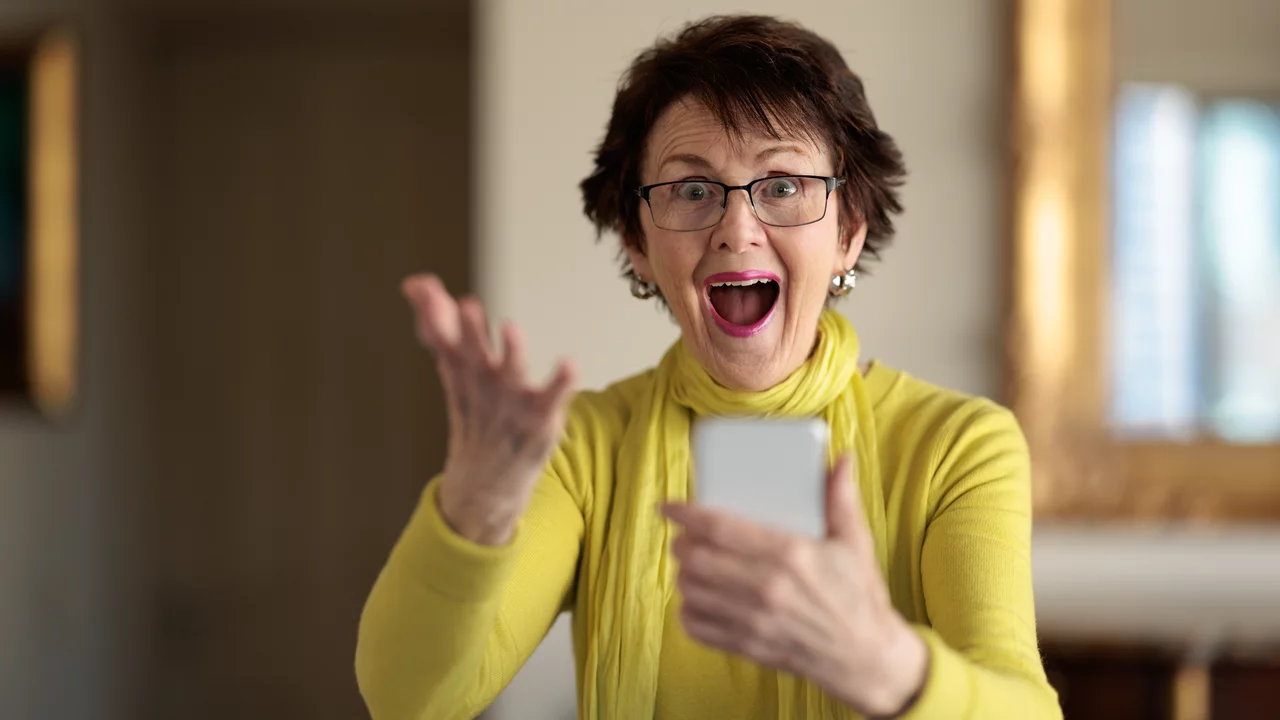 Eine fröhliche ältere Frau mit Brille trägt einen leuchtend gelben Pullover und einen Schal. Sie hält ein Smartphone in der Hand und zeigt mit der anderen Hand nach oben. Ihr Gesichtsausdruck drückt Überraschung und Freude aus.