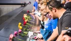 Menschen stehen an einem Mahnmal, während sie Blumen niederlegen und in Bücher schauen. Die Atmosphäre ist nachdenklich und respektvoll, mit mehreren Rosen, die entlang der Wand platziert sind.