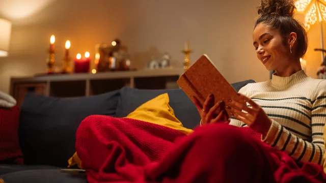 Eine Frau sitzt gemütlich auf einem Sofa, eingekuschelt in eine rote Decke, und liest ein Buch. Sie lächelt und befindet sich in einer warmen, einladenden Atmosphäre mit sanften Lichtquellen im Hintergrund.