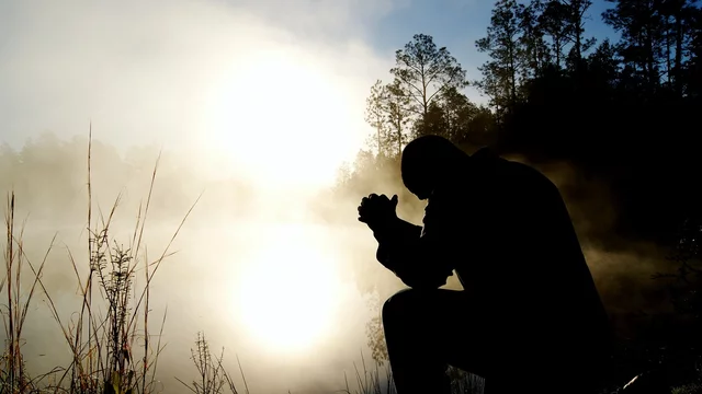 Eine silhouettierte Person kniet am Ufer eines ruhigen, nebligen Gewässers. Sie hält die Hände zusammen und blickt nach unten, während die Sonne im Hintergrund aufgeht und eine warme, goldene Lichtstimmung erzeugt. Umgeben von Bäumen und sanften Sträuchern.
