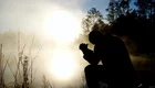 Eine silhouettierte Person kniet am Ufer eines ruhigen, nebligen Gewässers. Sie hält die Hände zusammen und blickt nach unten, während die Sonne im Hintergrund aufgeht und eine warme, goldene Lichtstimmung erzeugt. Umgeben von Bäumen und sanften Sträuchern.