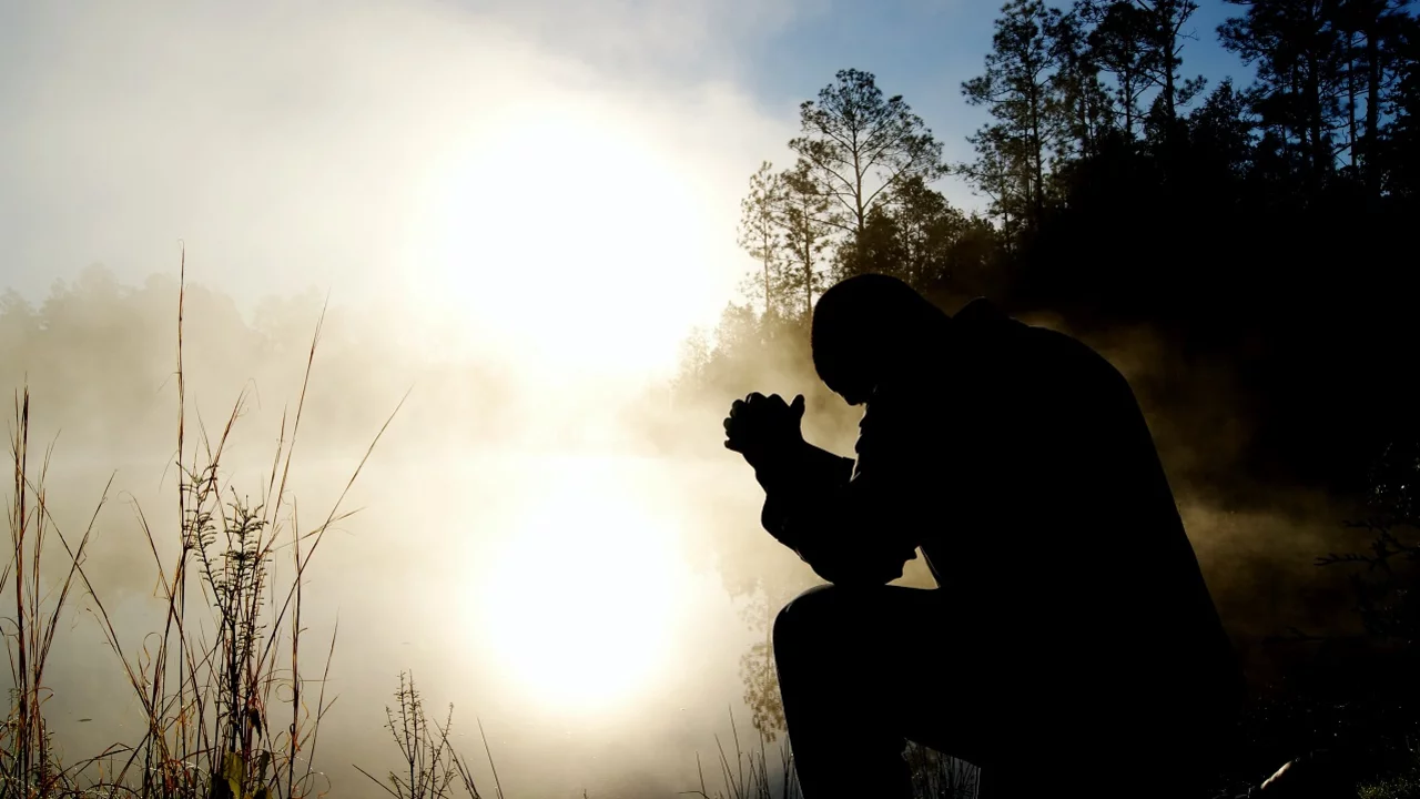 Eine silhouettierte Person kniet am Ufer eines ruhigen, nebligen Gewässers. Sie hält die Hände zusammen und blickt nach unten, während die Sonne im Hintergrund aufgeht und eine warme, goldene Lichtstimmung erzeugt. Umgeben von Bäumen und sanften Sträuchern.