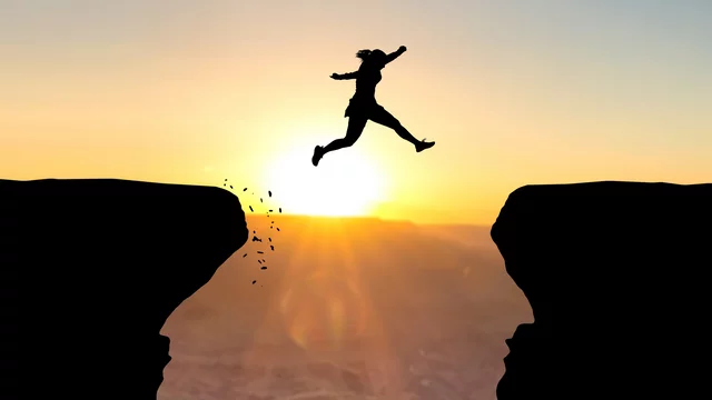 Eine Silhouette einer Person springt über eine Kluft, während die Sonne im Hintergrund aufgeht. Der Sprung symbolisiert den Übergang oder die Überwindung von Herausforderungen.