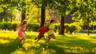 Drei Kinder rennen fröhlich über eine grüne Wiese im Sonnenschein. Die Mädchen tragen bunte Kleider, während der Junge in einem roten T-Shirt läuft. Bäume spenden Schatten und die Atmosphäre ist lebhaft und verspielt.