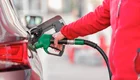 Eine Person in rotem Oberteil tankt ein Auto. Sie hält einen grünen Zapfhahn, der in den Tank des Fahrzeugs eingeführt ist. Im Hintergrund sind unscharfe Pumpen und eine Tankstelle sichtbar.