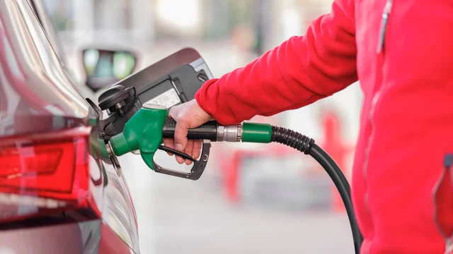 Eine Person in rotem Oberteil tankt ein Auto. Sie hält einen grünen Zapfhahn, der in den Tank des Fahrzeugs eingeführt ist. Im Hintergrund sind unscharfe Pumpen und eine Tankstelle sichtbar.