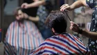 Ein Friseur schneidet einem Mann die Haare. Beide tragen gestreifte Umhänge, während der Friseur mit einer Schere arbeitet. Im Hintergrund ist ein weiterer Kunde zu sehen, der ebenfalls frisiert wird. Die Szene spielt in einem Barbershop mit entspannter Atmosphäre.