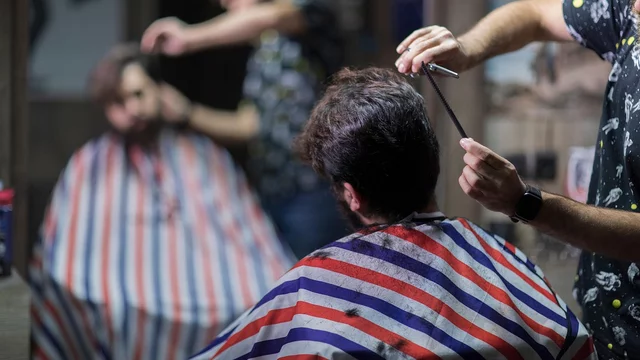 Ein Friseur schneidet einem Mann die Haare. Beide tragen gestreifte Umhänge, während der Friseur mit einer Schere arbeitet. Im Hintergrund ist ein weiterer Kunde zu sehen, der ebenfalls frisiert wird. Die Szene spielt in einem Barbershop mit entspannter Atmosphäre.