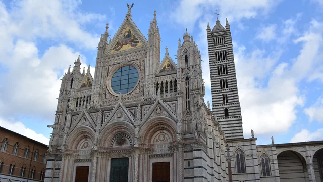 Die Kathedrale von Siena zeigt eine beeindruckende Fassade aus Marmor mit detaillierten Verzierungen und großen Fenstern. Neben der Kathedrale steht ein hoher Glockenturm, der die majestätische Architektur des Gebäudes unterstreicht. Der Himmel ist teils bewölkt.
