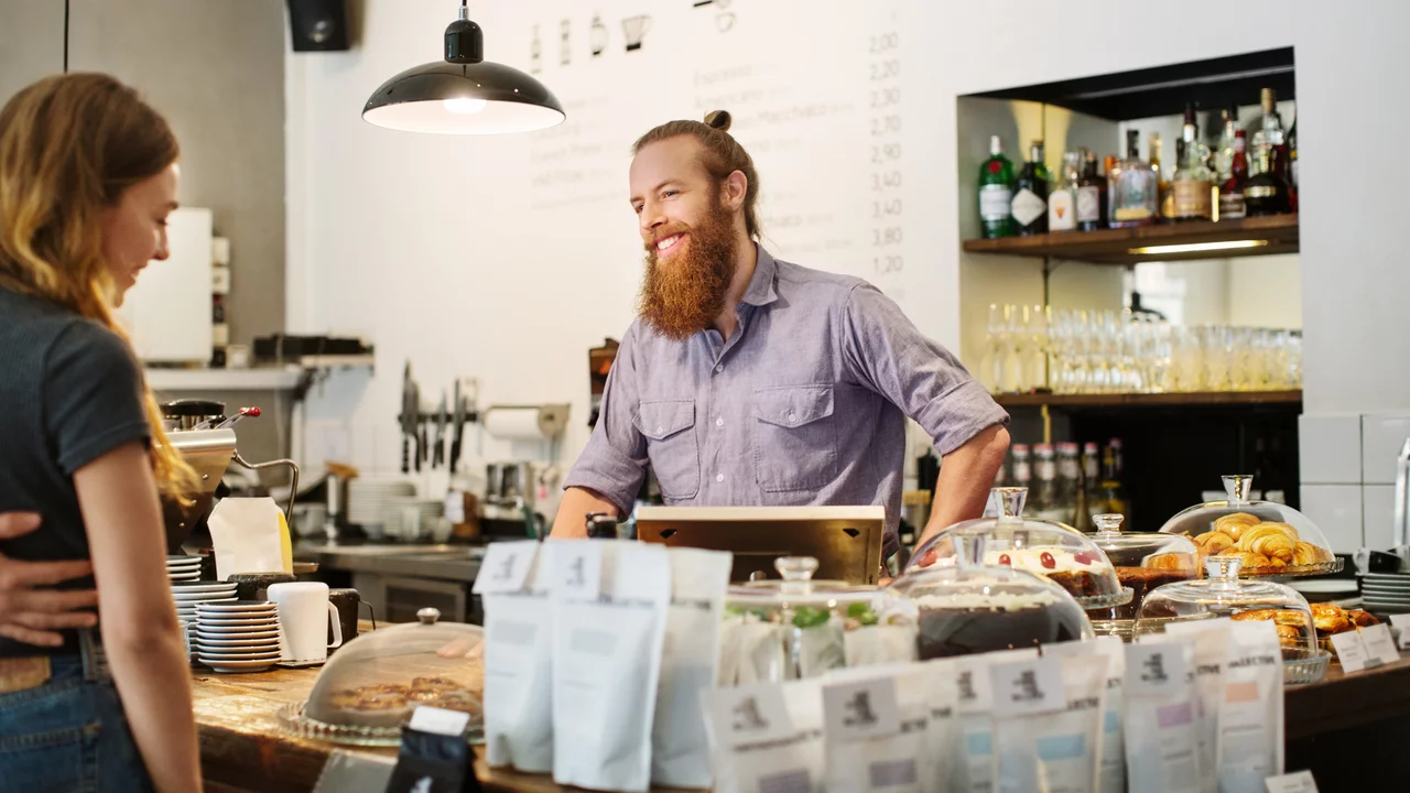 Ein Bartträger mit Hemd steht hinter einem Thekenbereich in einem Café. Er lächelt und kommuniziert mit einer Frau, die ihm gegenübersteht. Die Theke ist mit Kaffee, Snacks und verschiedenen Produkten dekoriert. Hintergrund zeigt Regale mit Getränken.