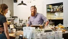 Ein Bartträger mit Hemd steht hinter einem Thekenbereich in einem Café. Er lächelt und kommuniziert mit einer Frau, die ihm gegenübersteht. Die Theke ist mit Kaffee, Snacks und verschiedenen Produkten dekoriert. Hintergrund zeigt Regale mit Getränken.