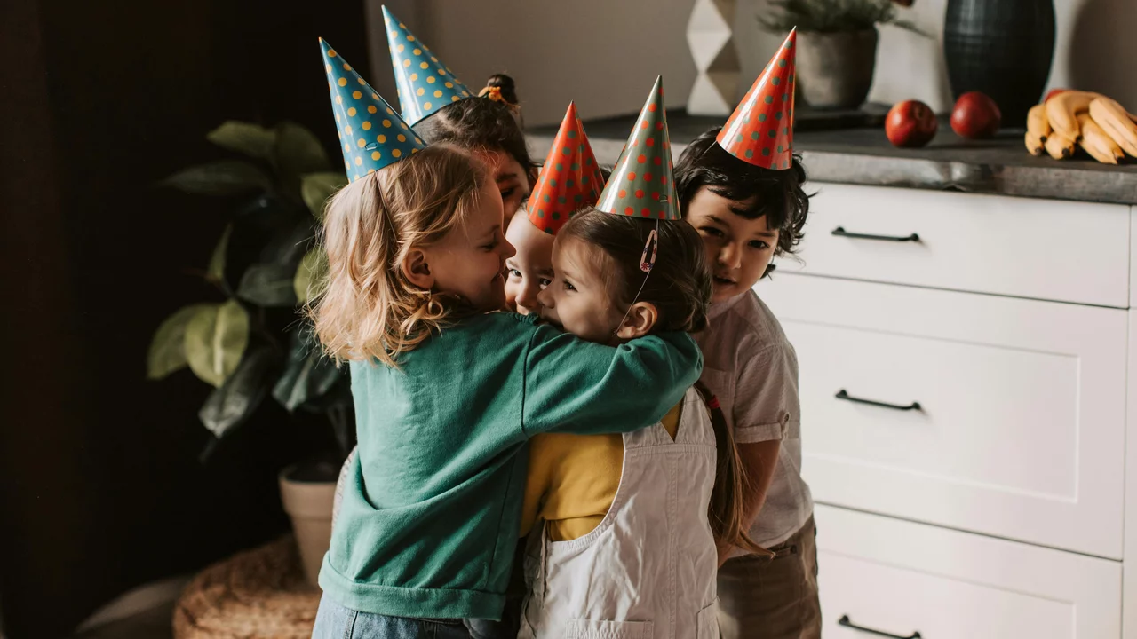 Vier Kinder umarmen sich fröhlich, während sie bunte Partyhüte tragen. Die Szene strahlt Freude und Freundschaft aus, im Hintergrund sind einige Äpfel und Bananen sichtbar. Die Atmosphäre wirkt warm und einladend, typisch für eine Feier.