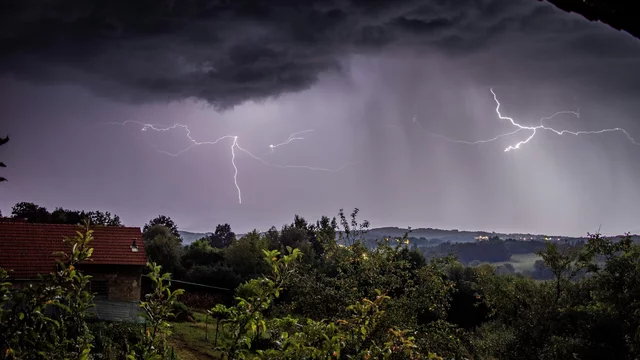 Blick aus dem Fenster in einen Baumbestandenen Garten und Hügellandschaft im Hintergrund. Dunkle Gewitterwolken und Blitze sind am Himmel zu sehen.