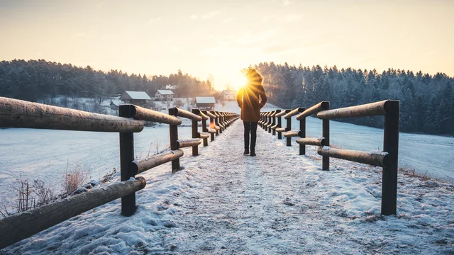 Eine Person geht auf einem schneebedeckten Weg, umgeben von Holzgeländern. Im Hintergrund ist eine Winterlandschaft mit Bäumen und einem Sonnenaufgang sichtbar, der das Szenario in warmes Licht taucht.
