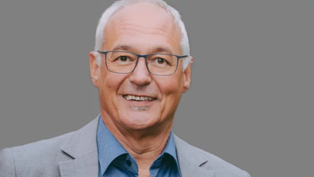 Ein lächelnder Mann mittleren Alters mit Brille, der einen hellgrauen Anzug und ein hellblaues Hemd trägt. Der Hintergrund ist grau. Der Mann schaut freundlich in die Kamera.