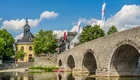 Eine historische Steinbrücke spannt sich über einen ruhigen Fluss, umgeben von grünen Bäumen und einem charmanten Gebäude. Im Hintergrund sind Flaggen sichtbar und der Himmel ist klar und sonnig, was eine ruhige, einladende Atmosphäre schafft.