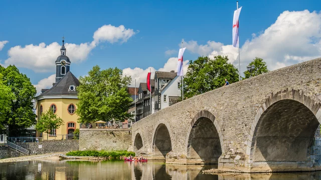Eine historische Steinbrücke spannt sich über einen ruhigen Fluss, umgeben von grünen Bäumen und einem charmanten Gebäude. Im Hintergrund sind Flaggen sichtbar und der Himmel ist klar und sonnig, was eine ruhige, einladende Atmosphäre schafft.