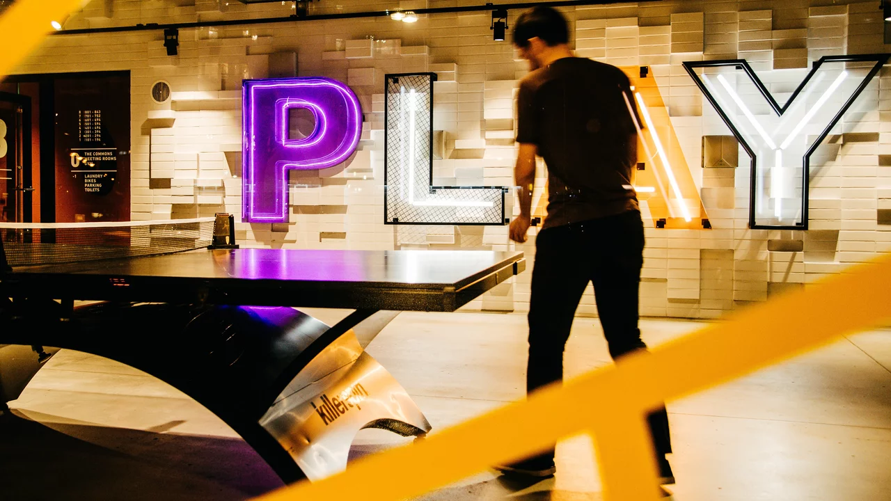 Ein Mann steht an einem Tisch, der möglicherweise für ein Spiel genutzt wird. Im Hintergrund sind große, beleuchtete Buchstaben zu sehen, die das Wort "PLAY" bilden. Die Beleuchtung ist farbenfroh und schafft eine spielerische Atmosphäre.