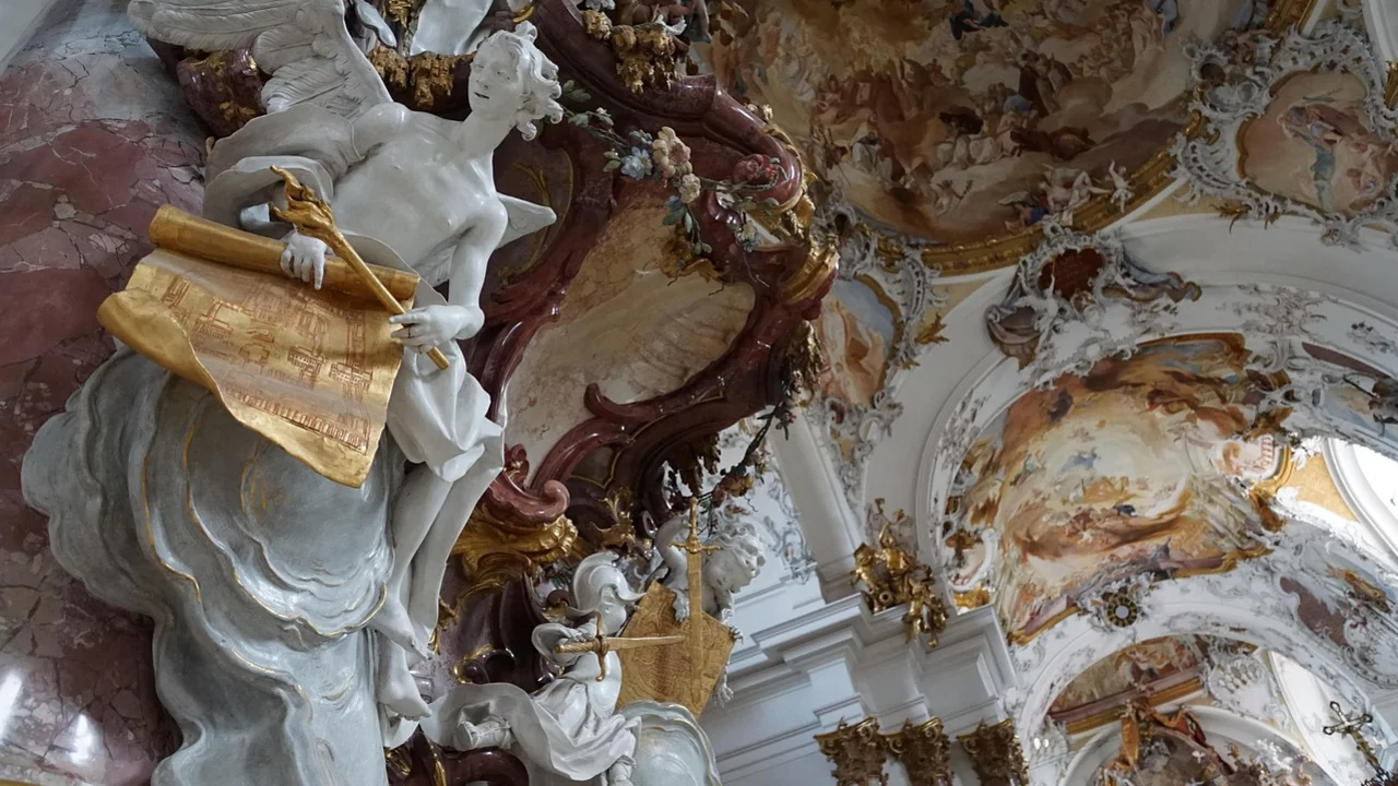 Eine barocke Wandgestaltung mit einer beeindruckenden Skulptur eines Engels, der Schriftrollen hält. Die Decke ist kunstvoll bemalt, mit religiösen Szenen und lebhaften Farben. Der gesamte Raum vermittelt einen Eindruck von Pracht und Spiritualität.