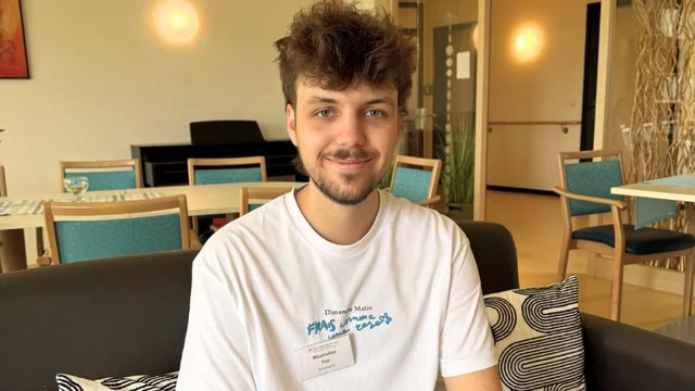 Ein junger Mann mit lockigem Haar sitzt auf einem Sofa in einem hellen Raum. Er trägt ein weißes T-Shirt mit einem Aufdruck und lächelt. Im Hintergrund sind Stühle und Tische zu sehen, die auf eine einladende Atmosphäre hinweisen.