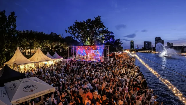 Ein großes Publikum versammelt sich an einem Ufer mit Blick auf eine Bühne, auf der ein Konzert stattfindet. Im Hintergrund sind beleuchtete Gebäude und ein Wasserspiel zu sehen. Der Himmel ist schätzungsweise dunkelblau und die Atmosphäre ist festlich und lebhaft.