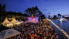 Ein großes Publikum versammelt sich an einem Ufer mit Blick auf eine Bühne, auf der ein Konzert stattfindet. Im Hintergrund sind beleuchtete Gebäude und ein Wasserspiel zu sehen. Der Himmel ist schätzungsweise dunkelblau und die Atmosphäre ist festlich und lebhaft.
